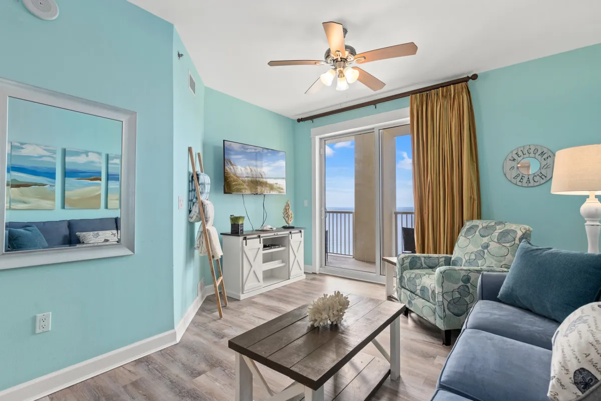 This inviting living room features light blue walls, beach-inspired decor, and a cozy seating area with a blue couch and patterned armchair, all oriented toward floor-to-ceiling balcony doors that open to breathtaking Gulf views. Natural sunlight fills the space, enhancing the airy, coastal vibe, while a wall-mounted flat screen TV adds a touch of modern convenience.