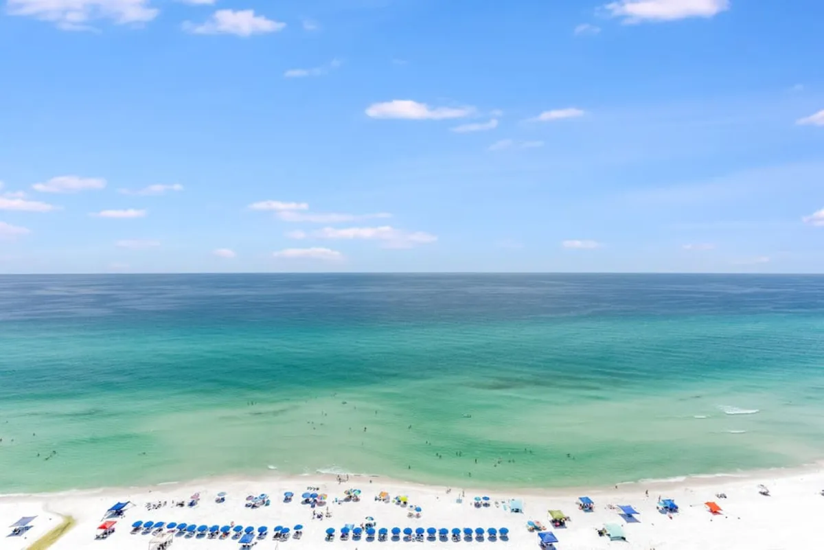 A sweeping view of the turquoise Gulf waters and white sandy beach lined with colorful umbrellas and lounge chairs. Small groups of people are seen enjoying the beach and water, conveying a lively yet relaxed atmosphere.