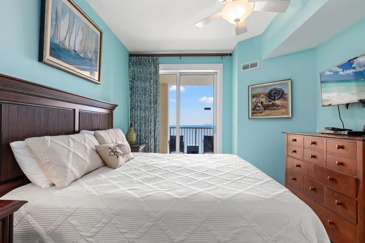 This image presents a spacious bedroom featuring a king-sized bed with crisp white bedding, a large wooden dresser topped by a flat-screen TV, and—most importantly—a glass balcony door offering a panoramic ocean view. The nautical-themed artwork and overall peaceful vibe reinforce the coastal ambiance, while the presence of the balcony visually connects the interior to the outdoors.