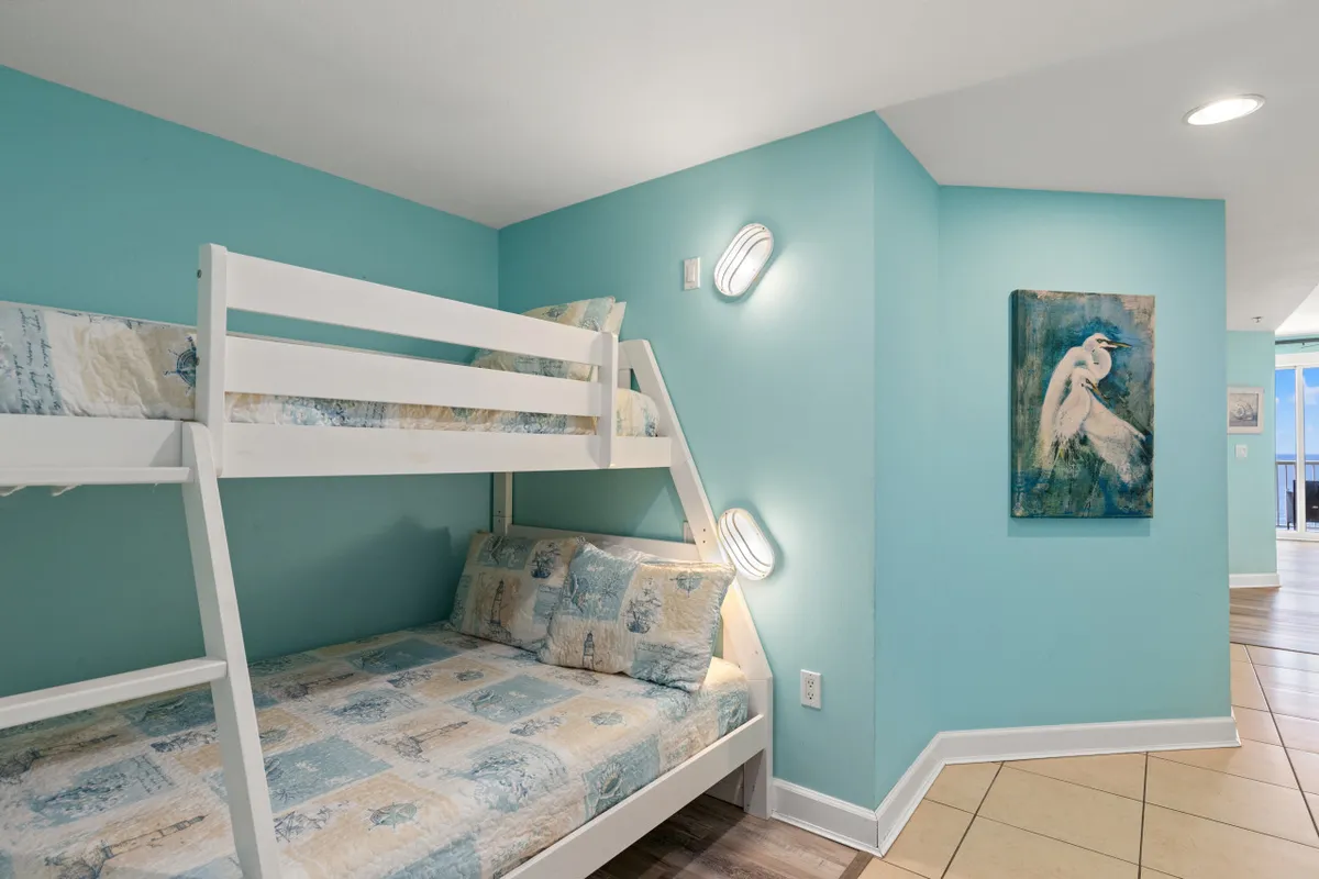A playful hallway bunk area with a white twin-over-full bunk bed, turquoise walls, nautical-themed bedding, and artwork. Wall-mounted lights provide cozy illumination, while the tiled floor leads toward an open area with a glimpse of a balcony view, evoking a sense of fun and togetherness.