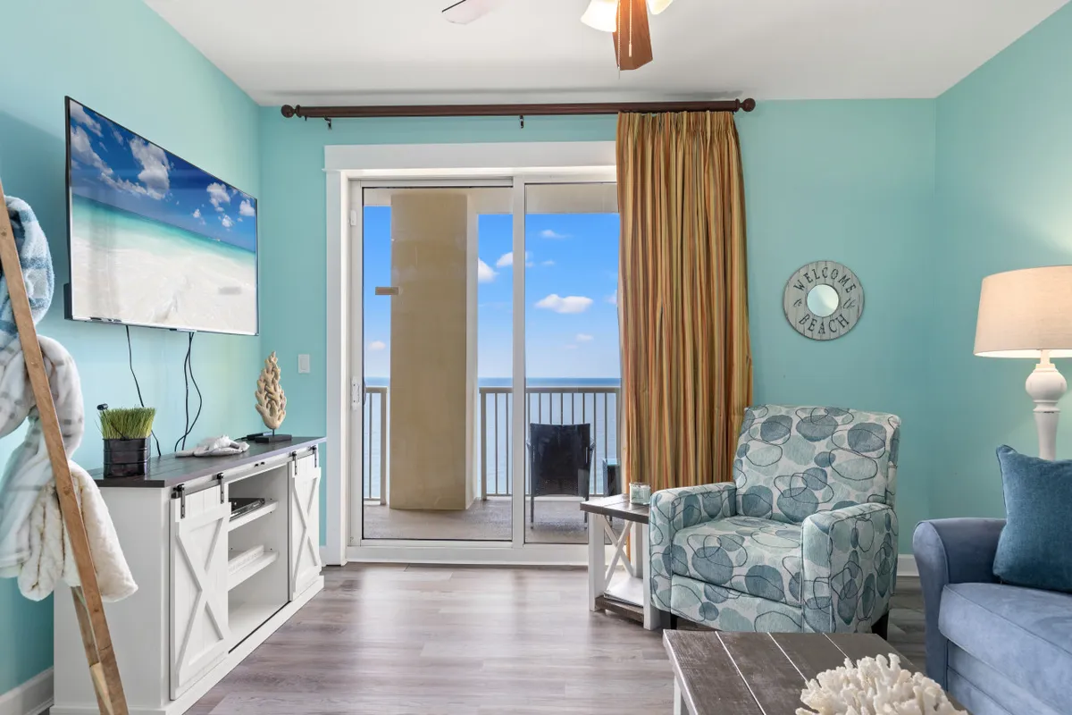 A bright, coastal-themed living room featuring a sliding glass door leading to a balcony with ocean views. The space includes a wall-mounted flat screen TV displaying a beach scene, a patterned armchair, and welcoming decor such as a 'Welcome Beach' sign.