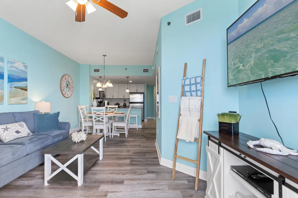 Showcasing a stylish, open-plan living area, this image features a blue accent wall with a mounted smart TV, a cozy light blue couch adorned with beach-themed cushions, and a dining area with white chairs and pendant lighting. The layout is bright, airy, and inviting, suggesting ample space for both work and leisure in a coastal-themed setting.