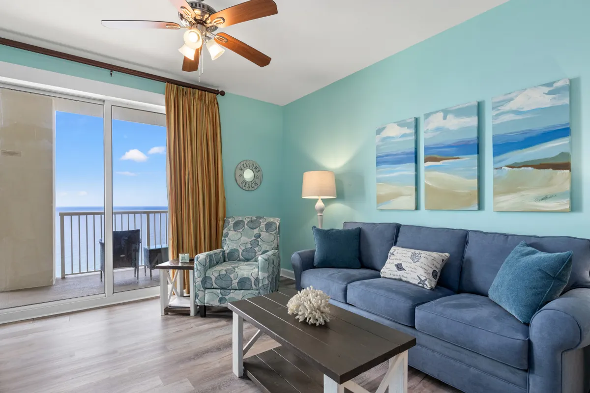 A cozy, inviting living room with a blue sofa adorned with sea-themed cushions, complemented by a three-panel beach landscape artwork above. The room features a patterned armchair with a side table and lamp, and a sliding glass door opens to a balcony with direct ocean views.