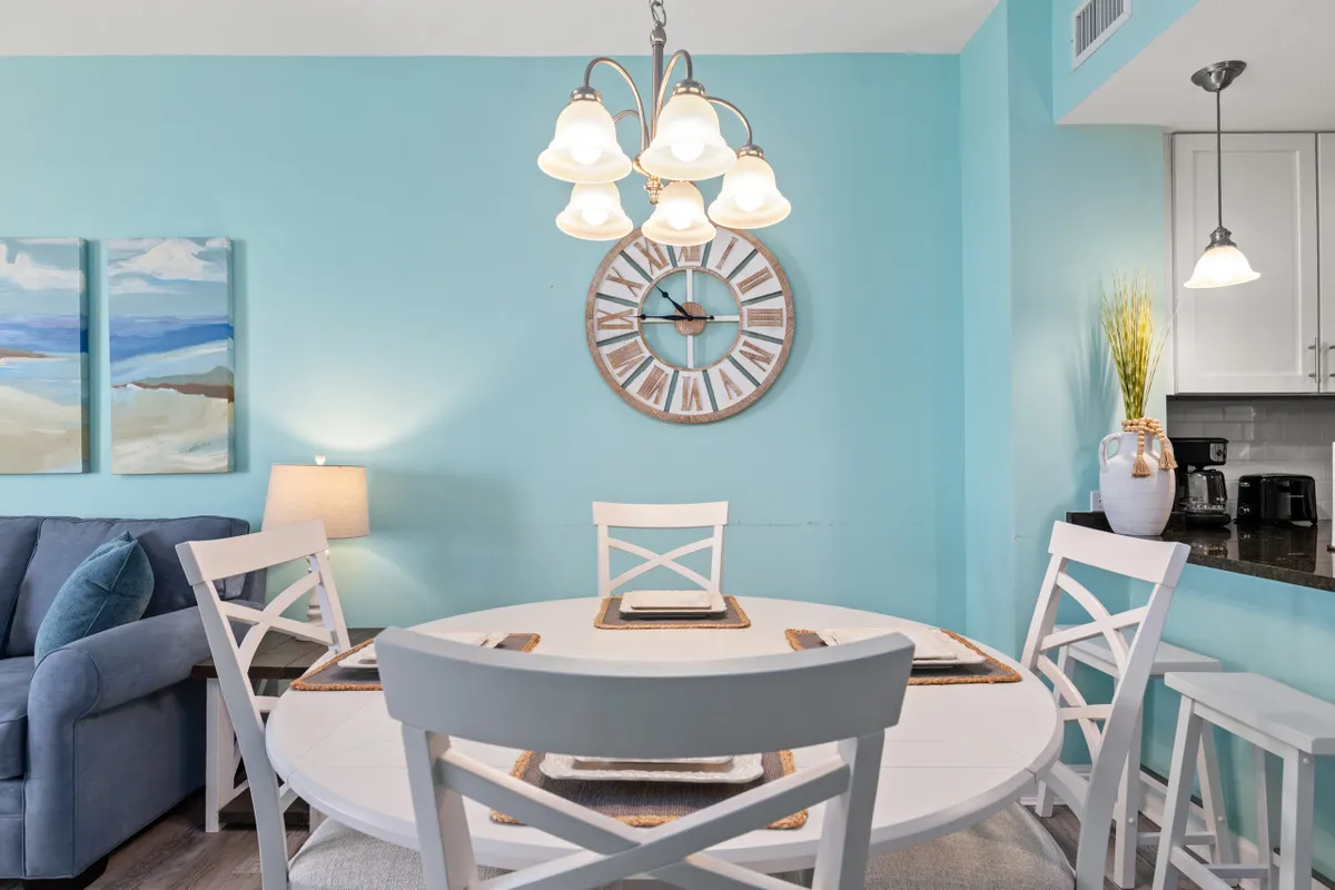 This image shows a round white dining table with placemats, a decorative wall clock, a blue accent wall, and an adjacent kitchen counter with bar stools. The setting is cheerful and welcoming, ideal for family meals or casual dining, and it visually connects the dining area to the kitchen.