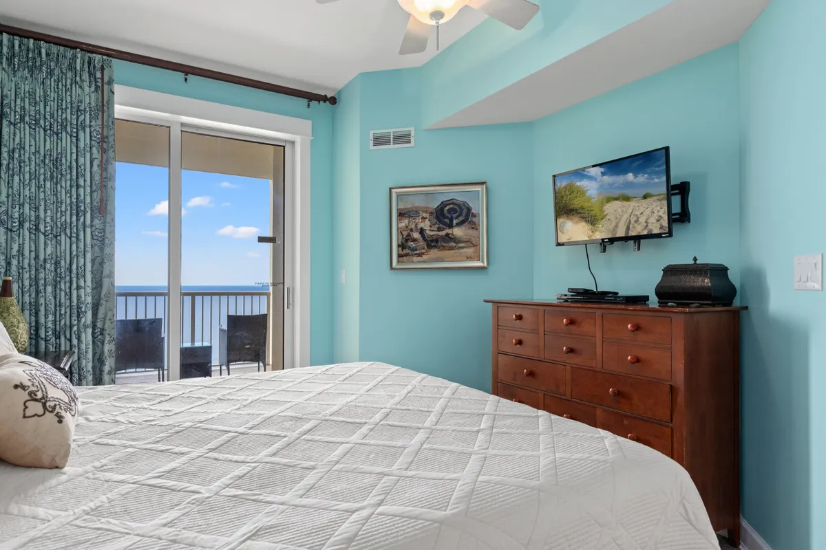 This image highlights a serene bedroom with a king bed, a mounted flat-screen TV, and a sliding glass door leading directly to a balcony overlooking the Gulf. The decor is airy and restful, while the balcony provides another visual anchor to the outdoors and opportunity for break-time relaxation.
