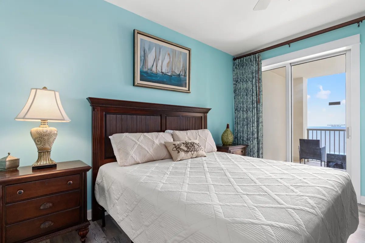 A tranquil primary bedroom featuring a king-sized bed with white quilted bedding, a large wooden headboard, and framed sailing boat artwork above the bed. Sunlight filters in through a balcony door that opens to an ocean view and outdoor seating, creating a peaceful, airy atmosphere ideal for relaxation and privacy.