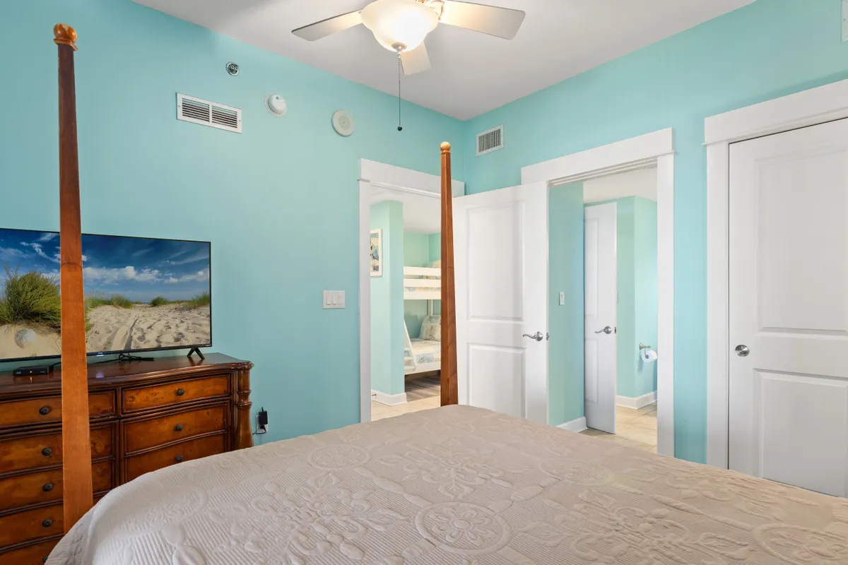 A family-friendly bedroom with a four-poster bed and beige quilted bedspread, a flat screen TV on a wooden dresser, and open doorways revealing adjoining rooms with bunk beds and a bathroom. The scene is bright, calm, and inviting, hinting at the proximity of different sleeping arrangements for families or groups.