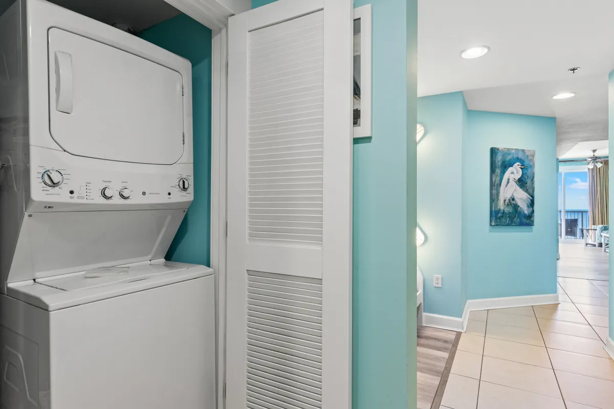 This image features a stacked washer and dryer unit against a turquoise accent wall, with a hallway leading to the living area and a glimpse of a balcony view. The decor is fresh and functional, emphasizing the convenience of in-unit laundry after beach outings.