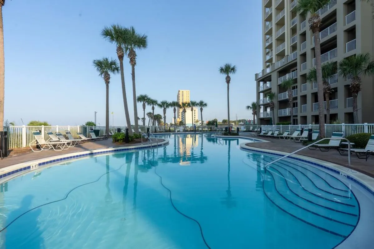 A large, curved swimming pool is at the center, surrounded by tall palm trees and a modern high-rise condo building. Poolside lounge chairs are arranged neatly, inviting relaxation and socializing in the sun-drenched tropical setting.