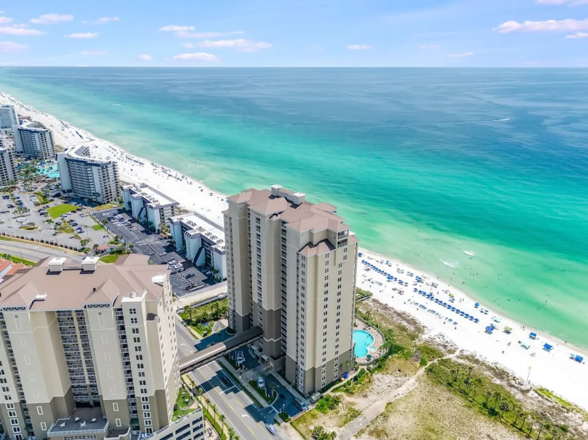 An exterior shot capturing the Grand Panama Tower 1 high-rise condo, adjacent community pool, white sandy beach, and umbrellas lining the shore. The scene is lively, with guests enjoying both pool and beach activities.