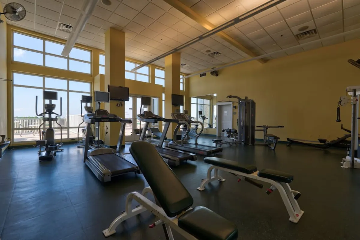 A modern on-site gym featuring treadmills, elliptical machines, weight benches, and multi-function equipment. Large windows offer glimpses of Gulf views, creating an energizing setting for vacation workouts and wellness routines.