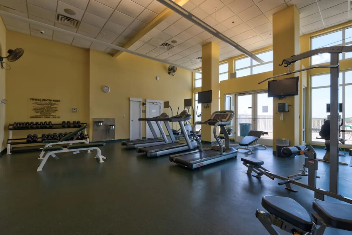 A modern fitness center with treadmills, a weight bench with dumbbells, a multifunctional weight machine, and large windows offering balcony views. The space is bright, airy, and equipped for a range of workouts.