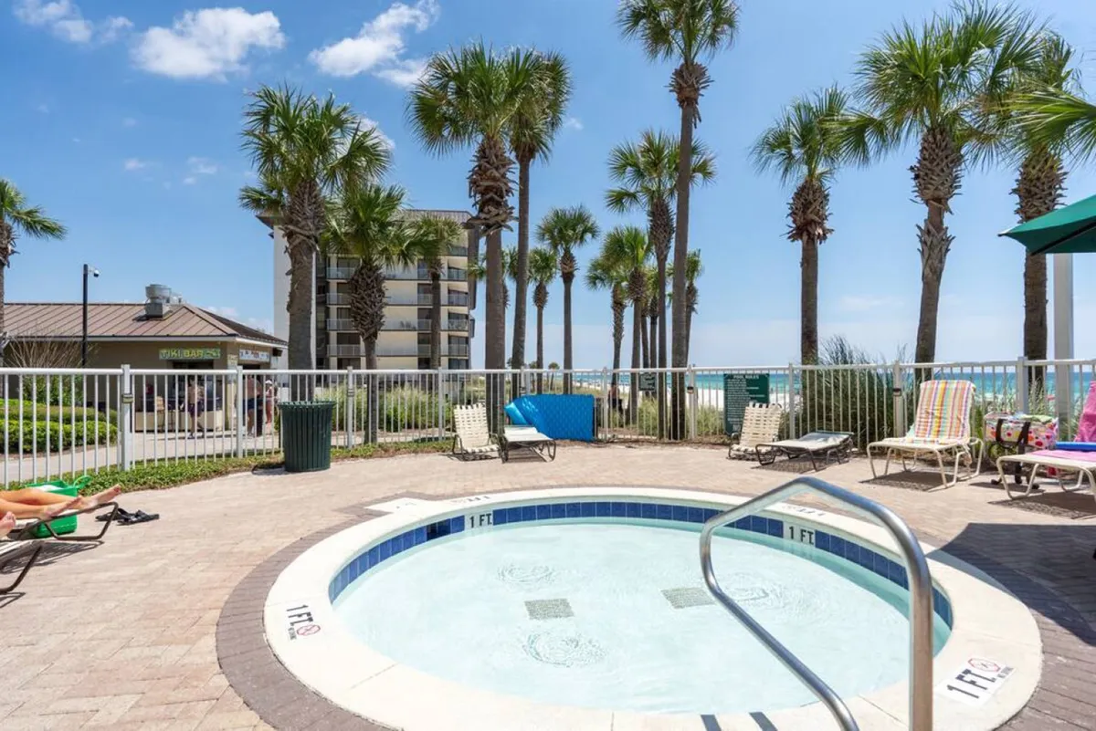 A family-friendly pool area encircled by sun loungers and tall palm trees, with a circular shallow pool featuring bubbling jets. A hint of the ocean is visible beyond the fencing, suggesting proximity to the beach.