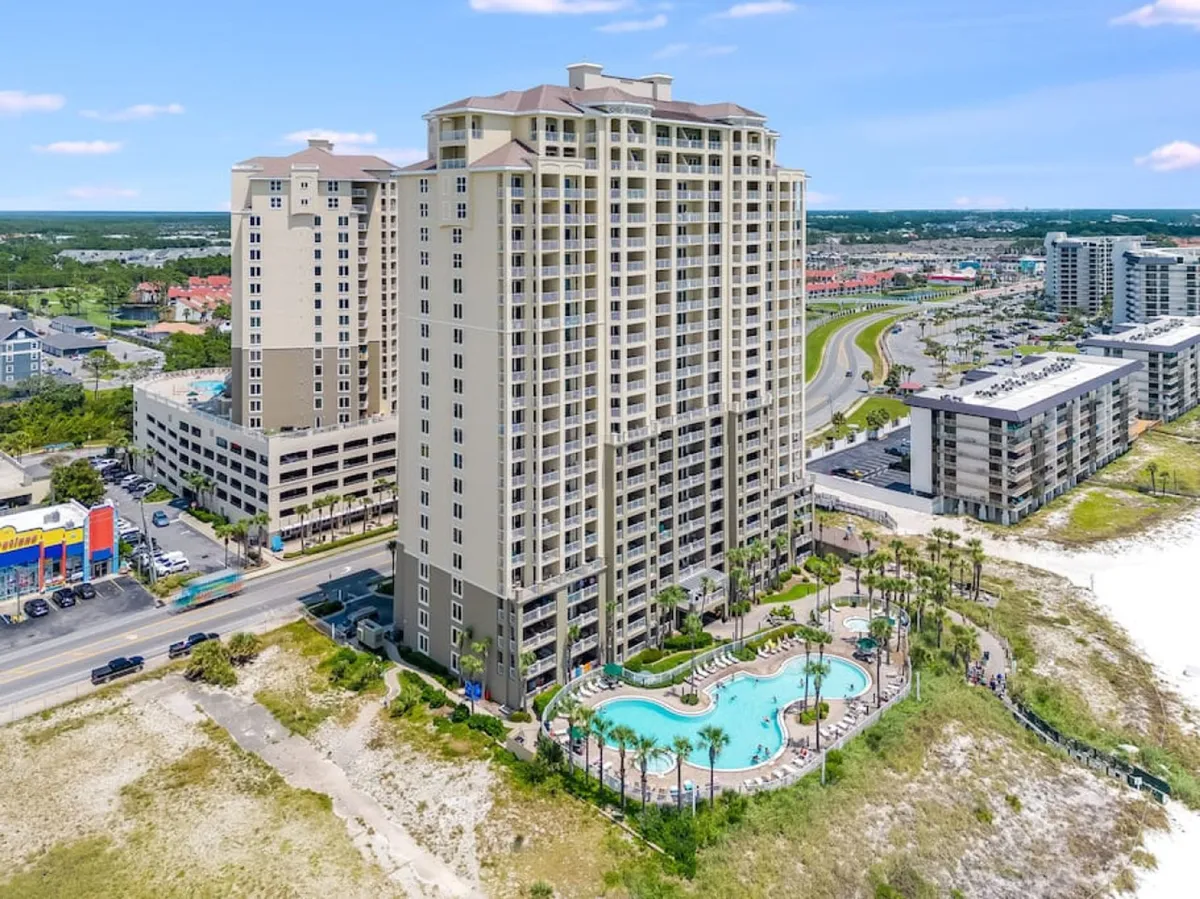 An exterior view of the Grand Panama Tower 1 condominium building, featuring a large, uniquely shaped community pool surrounded by lounge chairs, a sandy beachfront area adjacent to the pool, and the Gulf visible in the background. The image also captures the building’s proximity to the beach and nearby urban attractions.