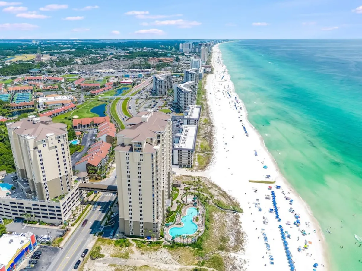 A sweeping exterior shot of high-rise beachfront condo buildings overlooking a sparkling community pool, with a wide sandy beach and the turquoise Gulf stretching to the horizon. Rows of umbrellas and beachgoers dot the scenery, showing the lively, accessible beach environment.