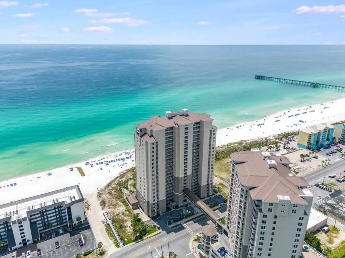 A wide-angle exterior view showing the Grand Panama Tower 1 beachfront condo building, the turquoise Gulf waters, and a pristine white sandy beach. The private walkway from the building leads directly onto the beach, with a pier visible in the distance.