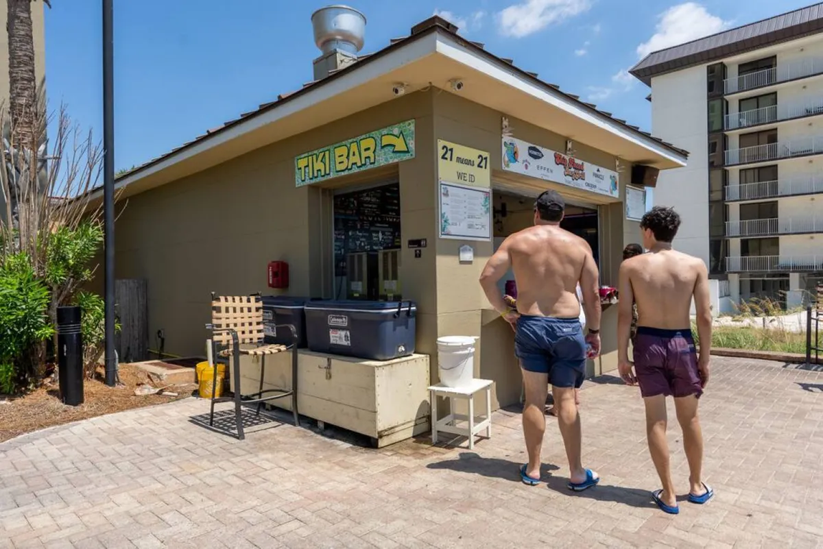 An exterior view of the resort’s Tiki Bar with signage, where two guests in swimwear order drinks. Outdoor seating and palm landscaping create a vibrant, social scene, with the high-rise building visible nearby.
