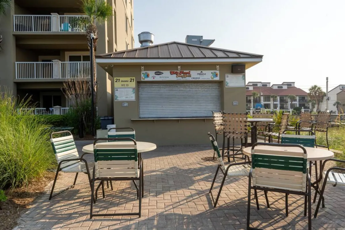 An exterior scene featuring a closed tiki bar with clear signage, outdoor seating, palm trees, and nearby resort buildings, all set in a landscaped communal area adjacent to beachside amenities.