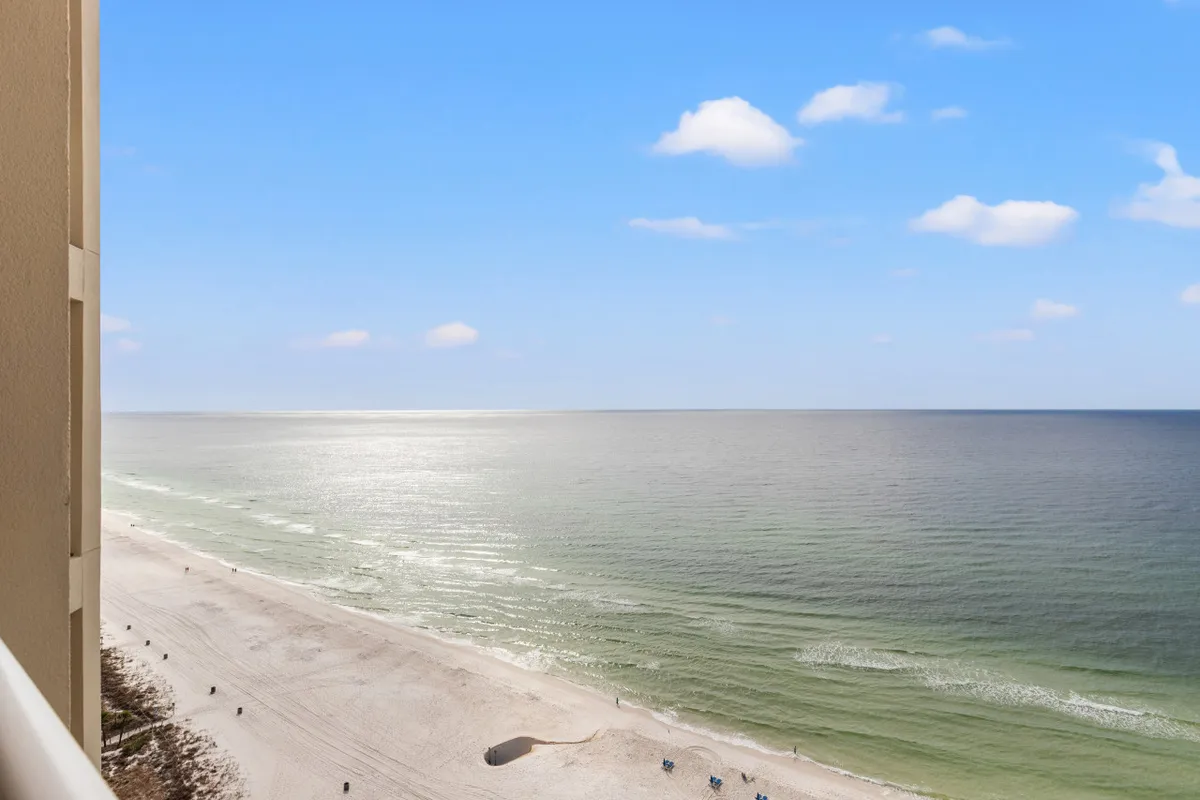 A high-floor balcony view overlooking the vast Gulf of Mexico, pristine white sand beach, and blue sky scattered with clouds. The balcony railing and part of the building’s facade are visible, emphasizing the condo’s direct beachfront perspective.