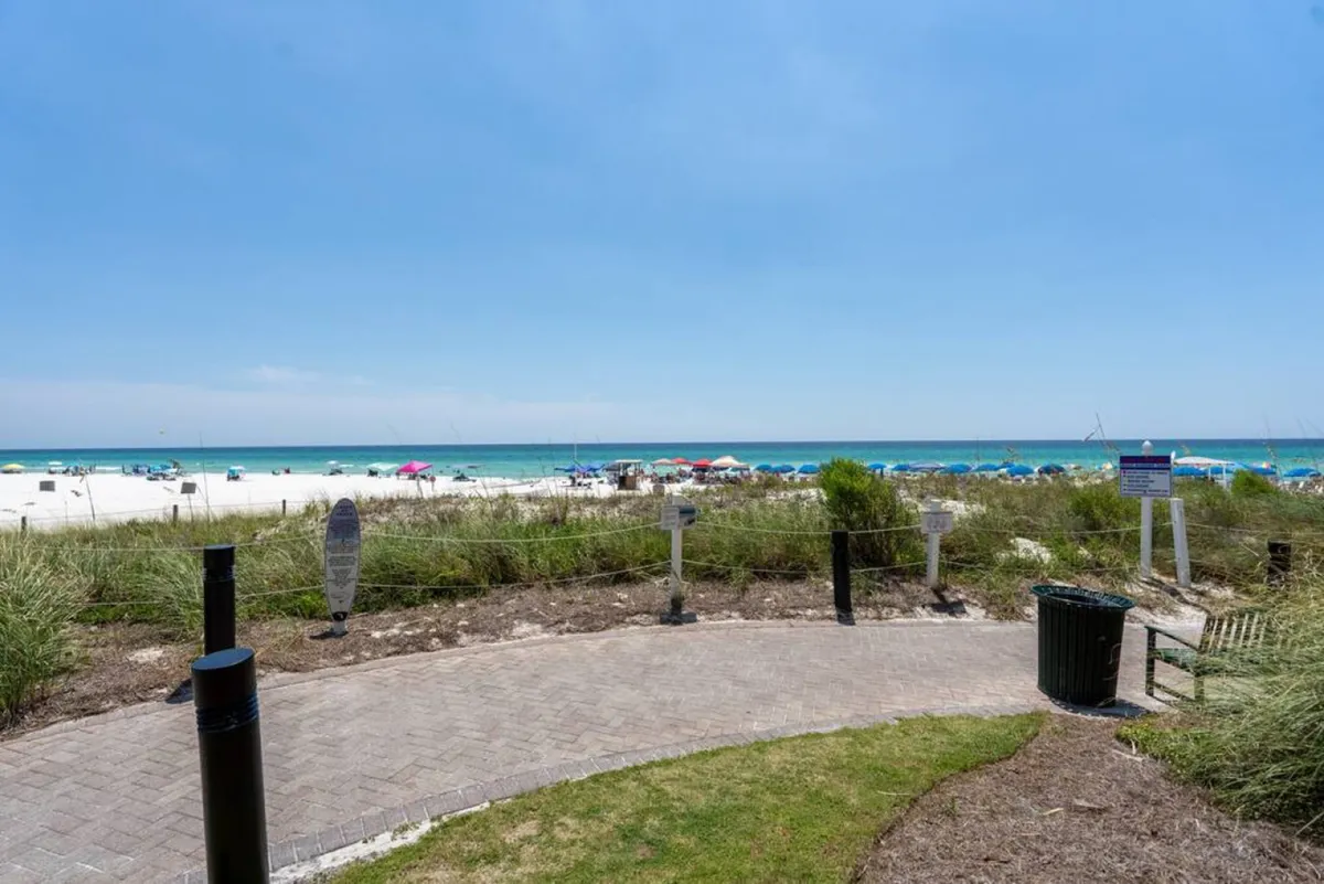 A vibrant exterior shot capturing the expansive white sandy beach of Panama City Beach, dotted with colorful umbrellas and bordered by turquoise Gulf waters. The paved walkway along the dunes and beach grass invites guests for strolls, with signage and amenities visible, setting the scene for water sports, sunbathing, and exploration.