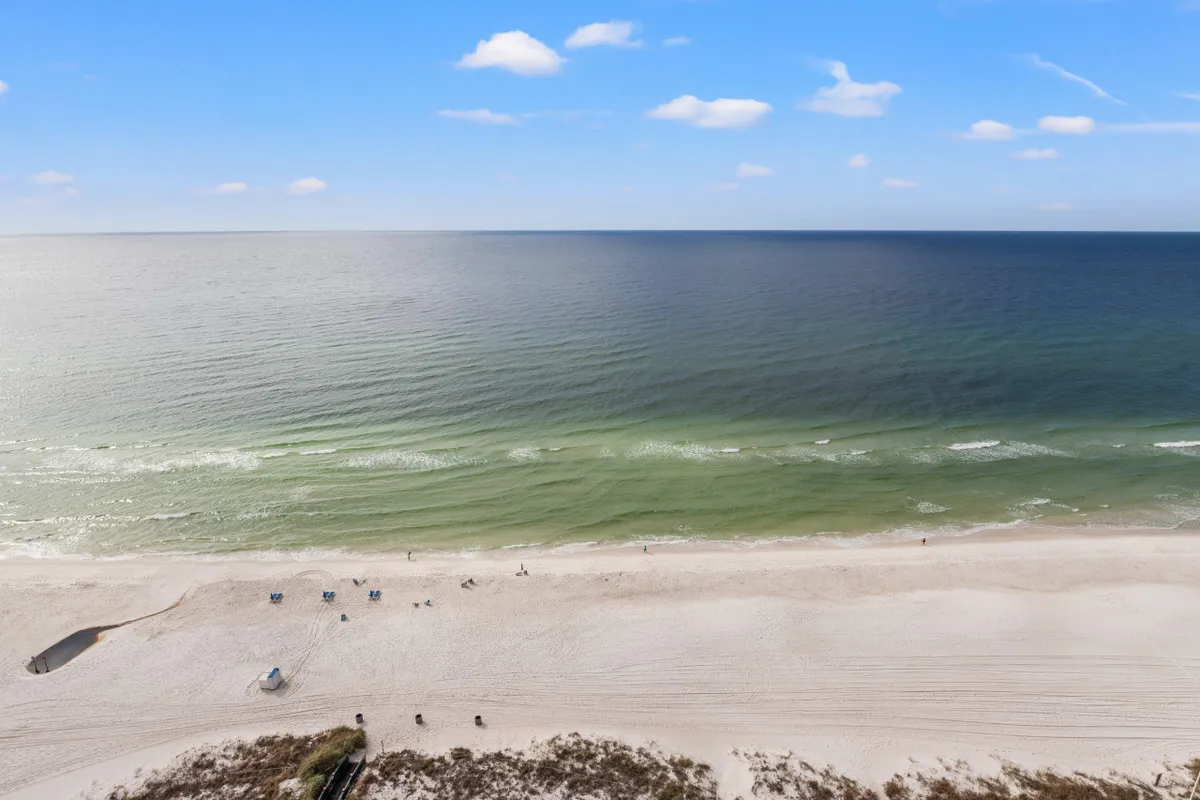 A wide view of the nearly empty sandy beach with gentle Gulf waves rolling ashore, a few beach chairs and an umbrella set up, and the vast ocean horizon stretching under a bright sky.